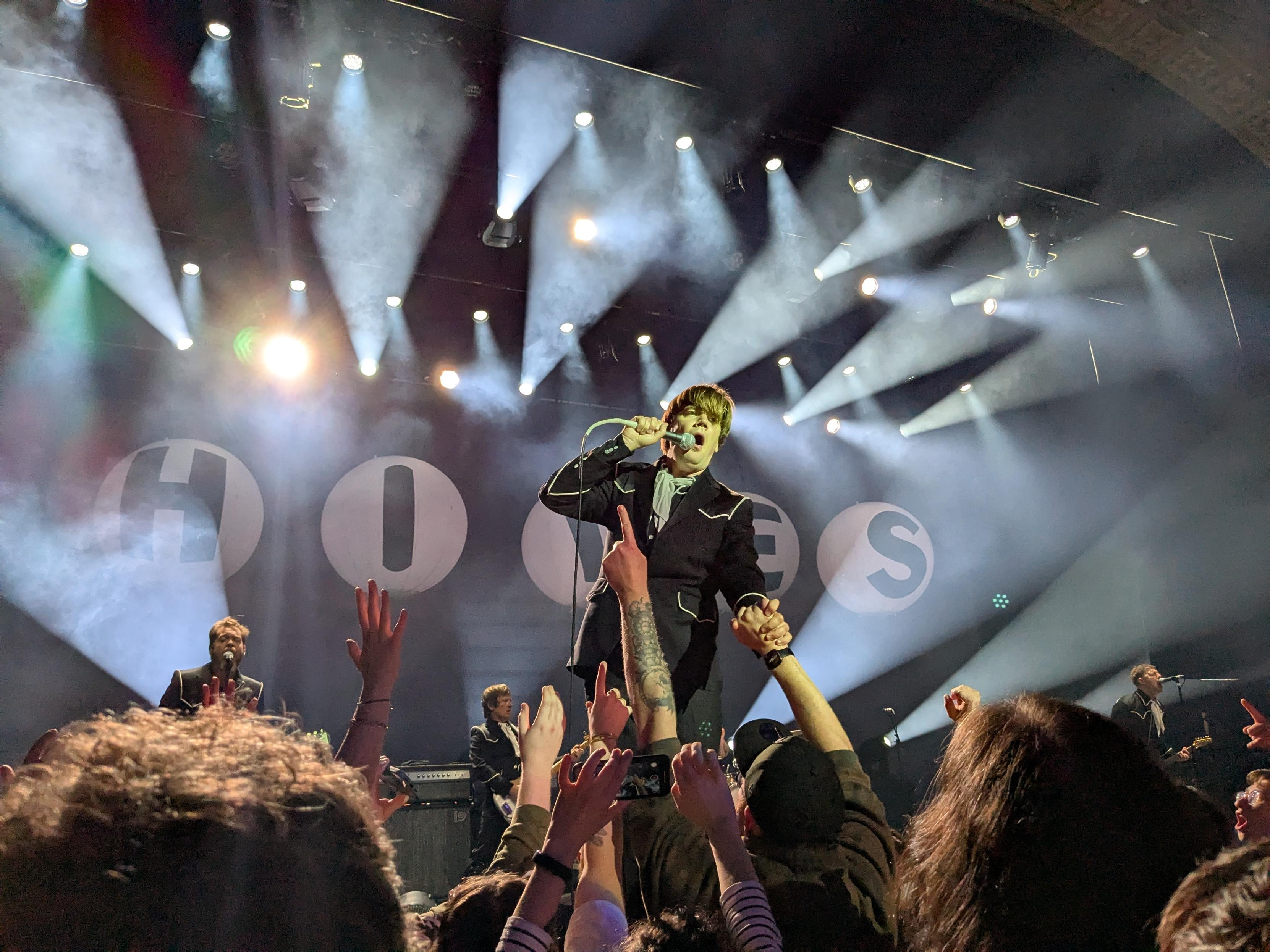 Lead singer of the hives up on stage leaning over the audience while hands reach up for him.  he is backlit by stage lights and it all looks very rock and roll